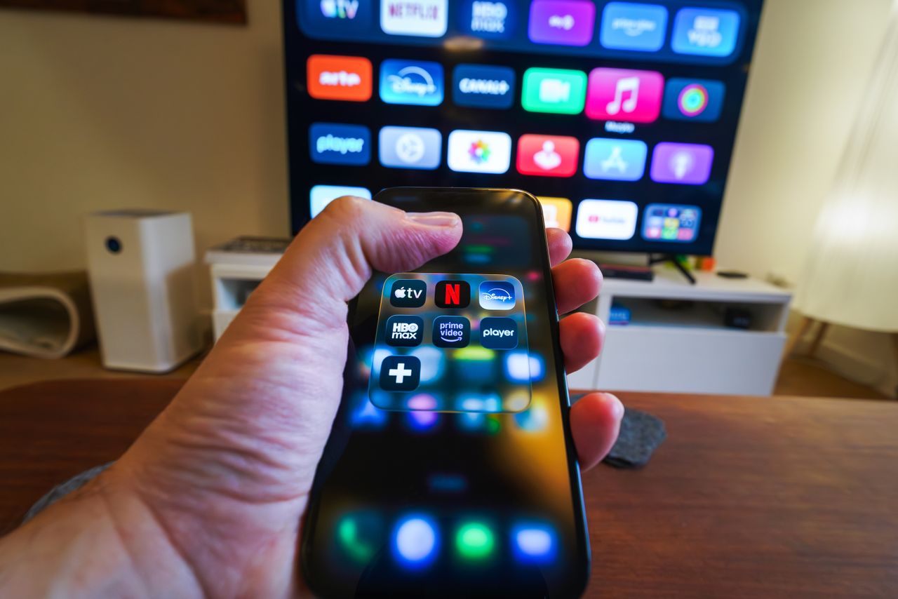 Services Close-up of a hand holding a phone displaying streaming apps in front of a TV with multiple app icons.
